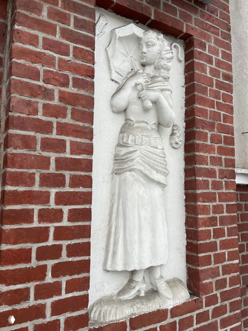 Барельеф женской фигуры с элементами драпировки и украшений; белая скульптура в глубокой нише кирпичного фасада, видны следы реставрации.