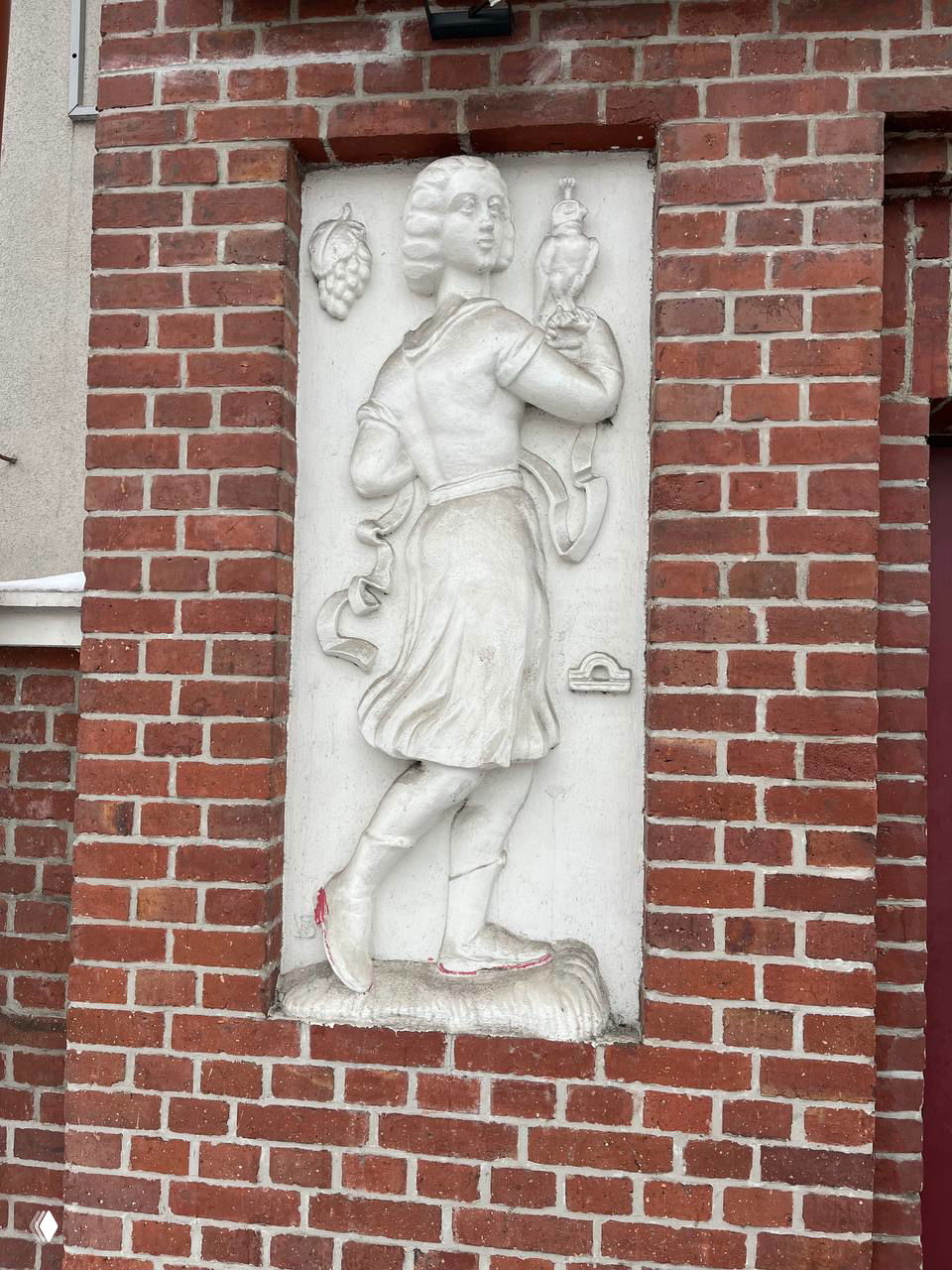 Барельеф девушки с гроздью винограда и атрибутами, рядом виден знак Весы; белая рельефная композиция на фоне кирпича у портала.