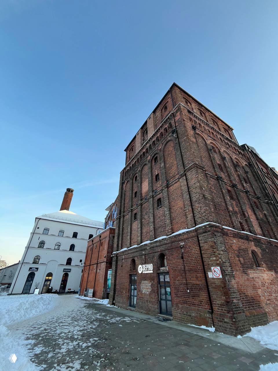 Угловой вид массивных кирпичных корпусов бывшего пивзавода, белый пристроенный корпус и высокая дымовая труба на морозном фоне.