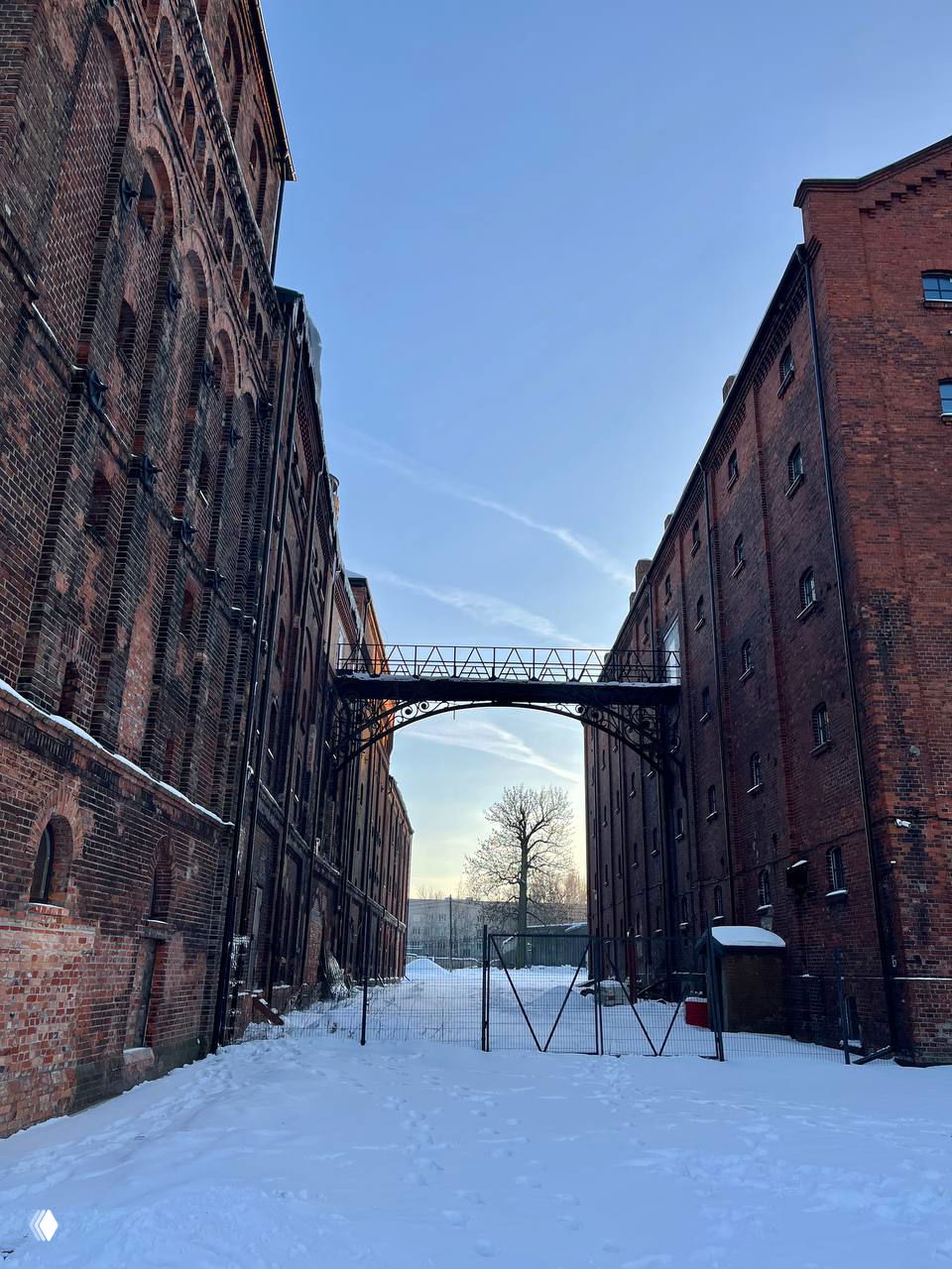 Проход между двумя кирпичными корпусами завода с подвесным мостиком над заснеженным двором, зимний свет и сплошной голубой небосвод.