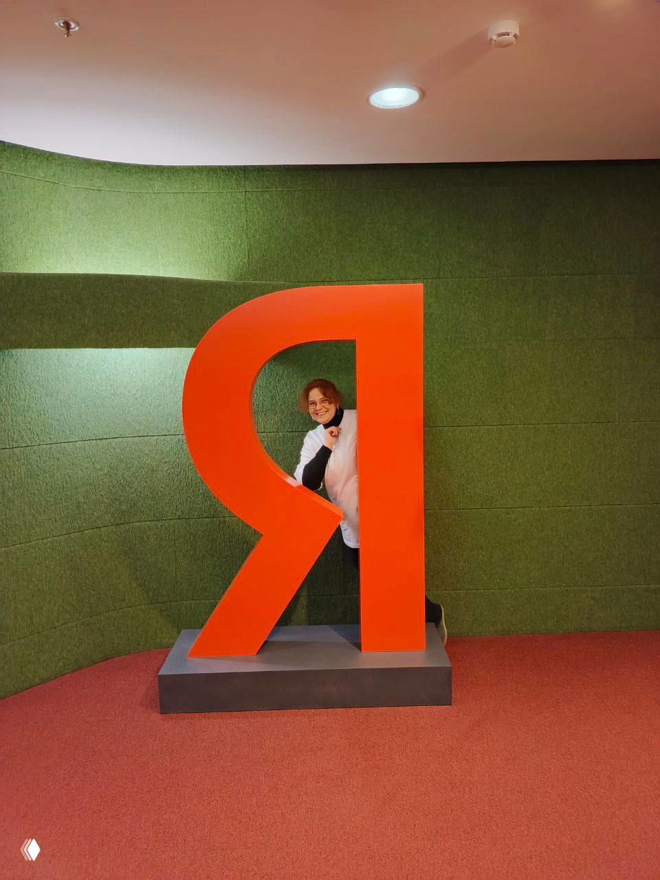 Женщина позирует внутри большой оранжевой буквы «Я» на фоне зелёной текстурной стены и красного коврового покрытия в офисе Яндекса.