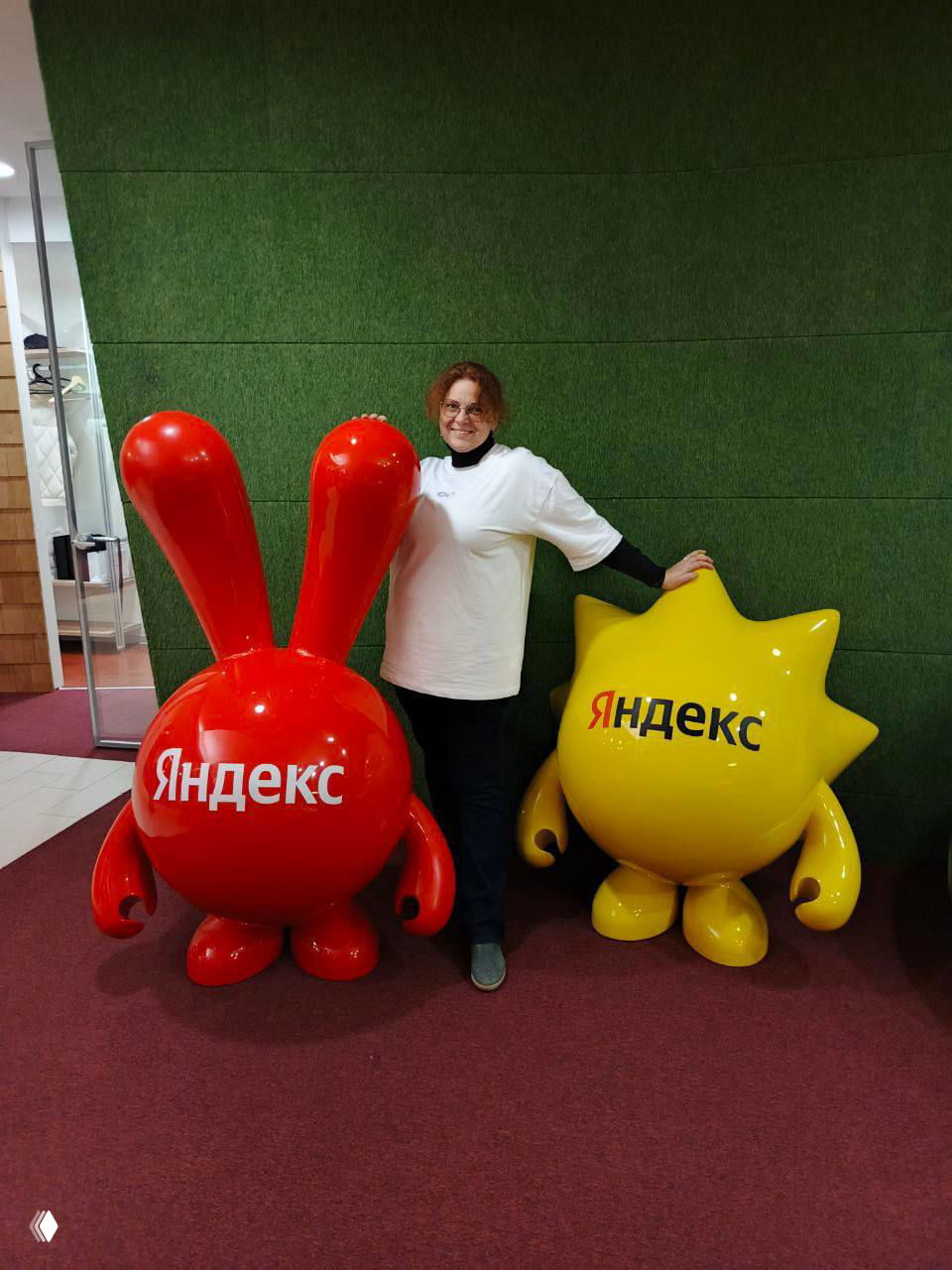 Женщина стоит между двумя крупными фигурками-талисманами Яндекса — красной и жёлтой — на фоне зелёной стены в офисном пространстве.