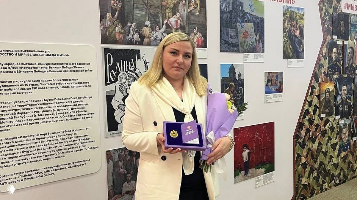 Елена Гроздева в светлом пальто держит футляр с почётным знаком и букет на фоне выставки с картинами — момент вручения награды