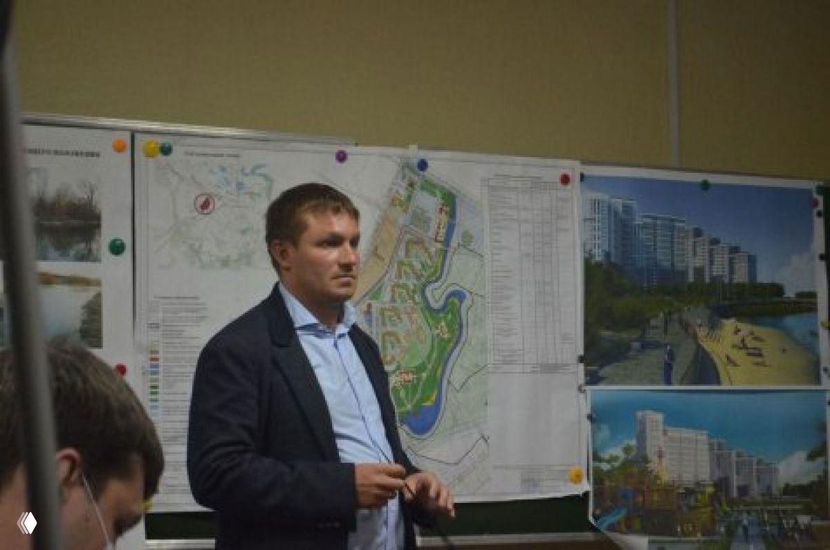 Мужчина в деловом костюме стоит на фоне архитектурных планов и визуализаций — презентация проектов застройки и городской инфраструктуры Рязанской области.