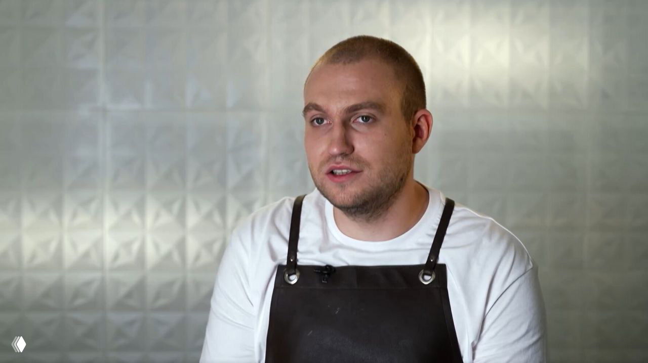 Портрет рязанского су‑шефа Егора Жукова в тёмном кожаном фартуке и белой футболке на нейтральном студийном фоне во время съёмок телевизионного проекта.