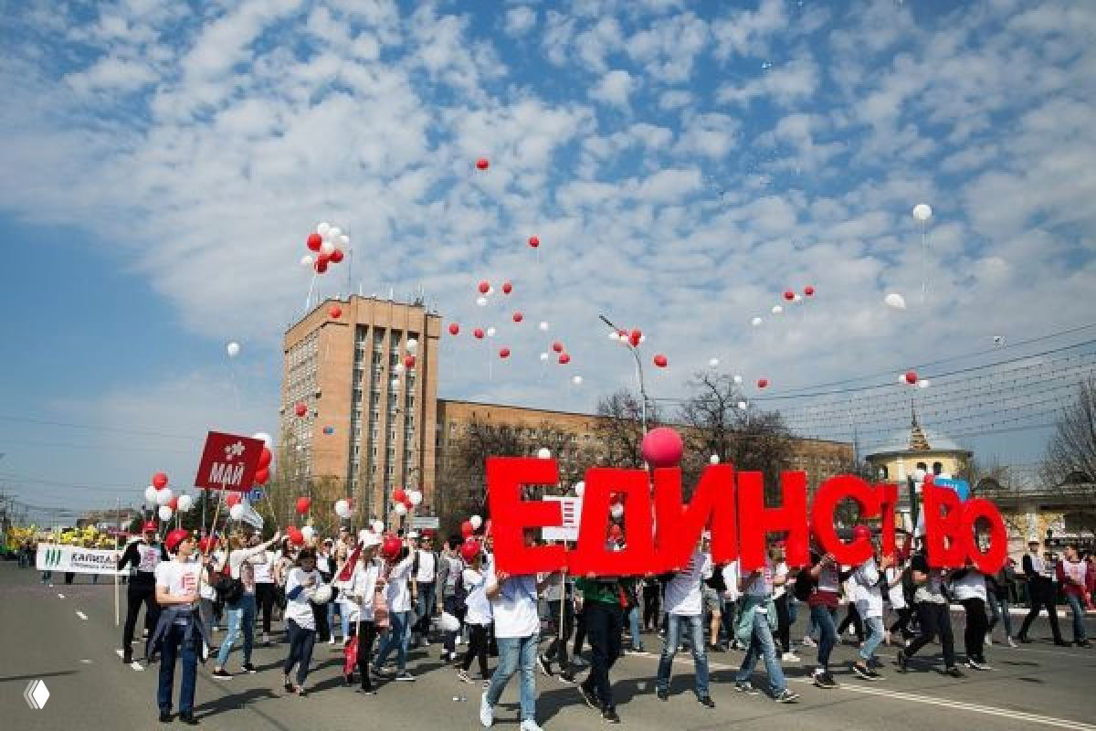 Шествие в городе с большой красной инсталляцией «Единство», участники с бело-красными шарами проходят мимо многоквартирных зданий и ясного неба в качестве фона.