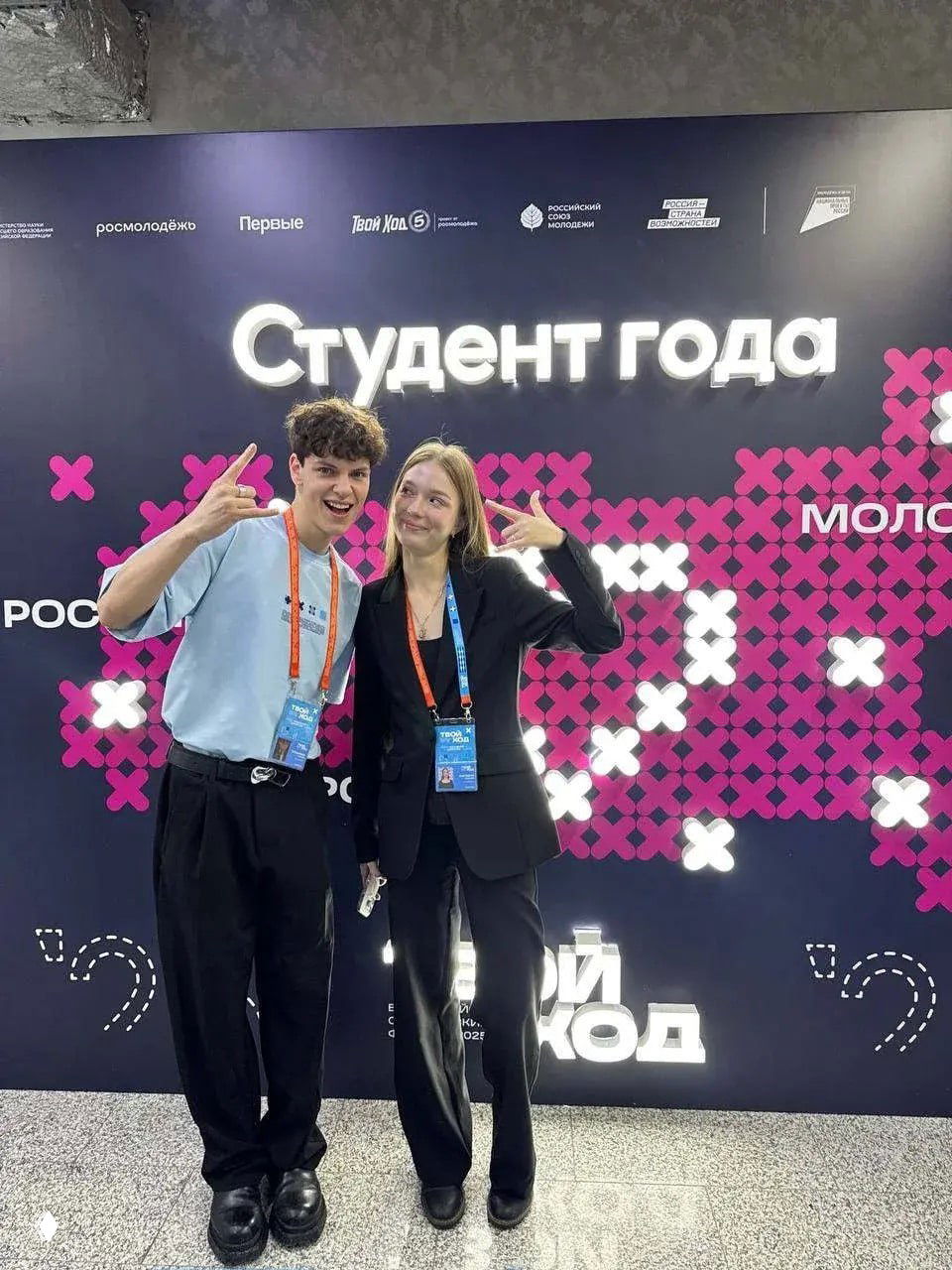 Два студента из Рязанского медколледжа на фоне баннера «Студент года» в Москве, с бейджами и радостными жестикуляциями