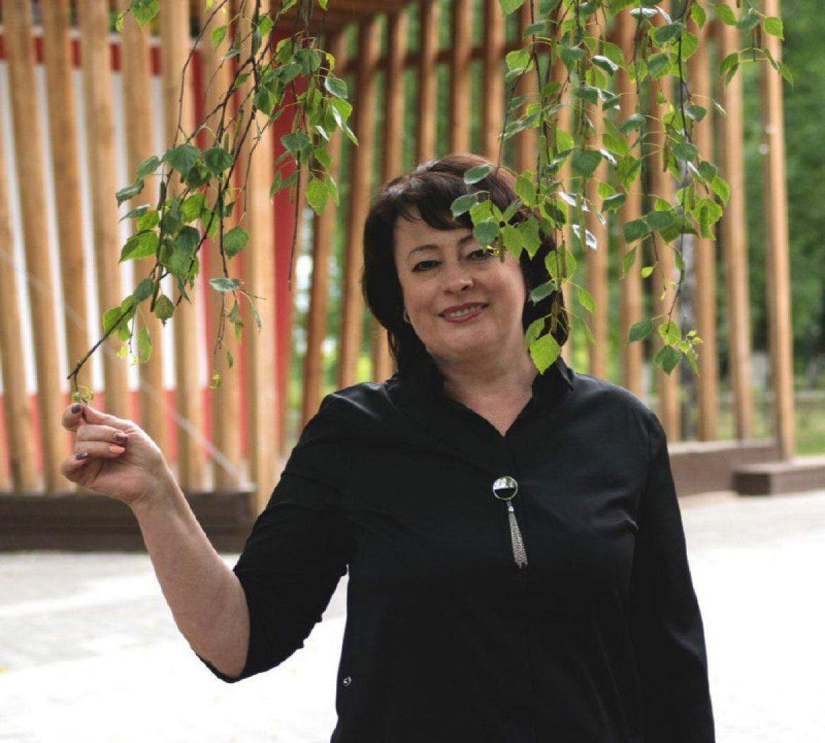 Портрет Евгении Рубцовой: улыбающаяся женщина средних лет на улице, держит ветку берёзы на фоне деревянных конструкций и парковой зоны.