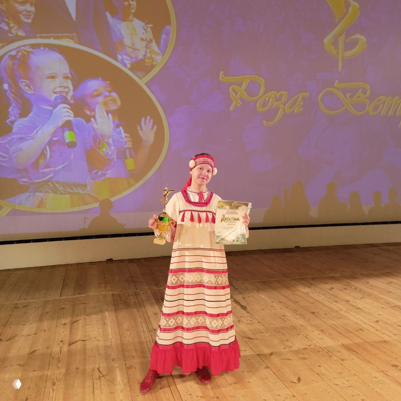 Солистка ансамбля Ева Орлова в традиционном платье держит кубок и диплом на деревянной сцене на фоне проекции мероприятия «Роза ветров»