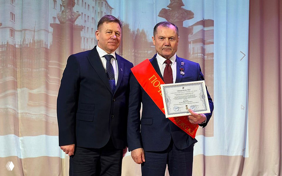 Двое мужчин в деловых костюмах на сцене во время церемонии: Игорь Мурог в красной ленте держит свидетельство о присвоении звания почётного гражданина Ряжска.