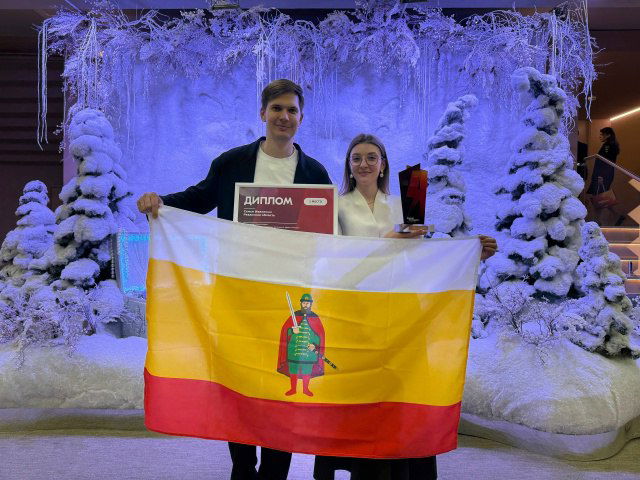 Мария и Никита Ивахины держат флаг Рязанской области и дипломы на фоне зимнего оформления после награждения в номинации «Семья молодых».