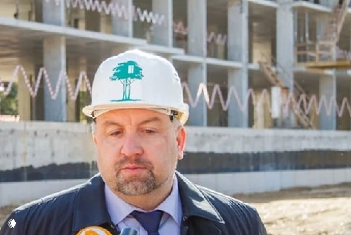 Мужчина в белой строительной каске с логотипом компании «Зелёный сад» на фоне недостроенного многоквартирного дома и строительных лесов — стройплощадка в Рязанской области.