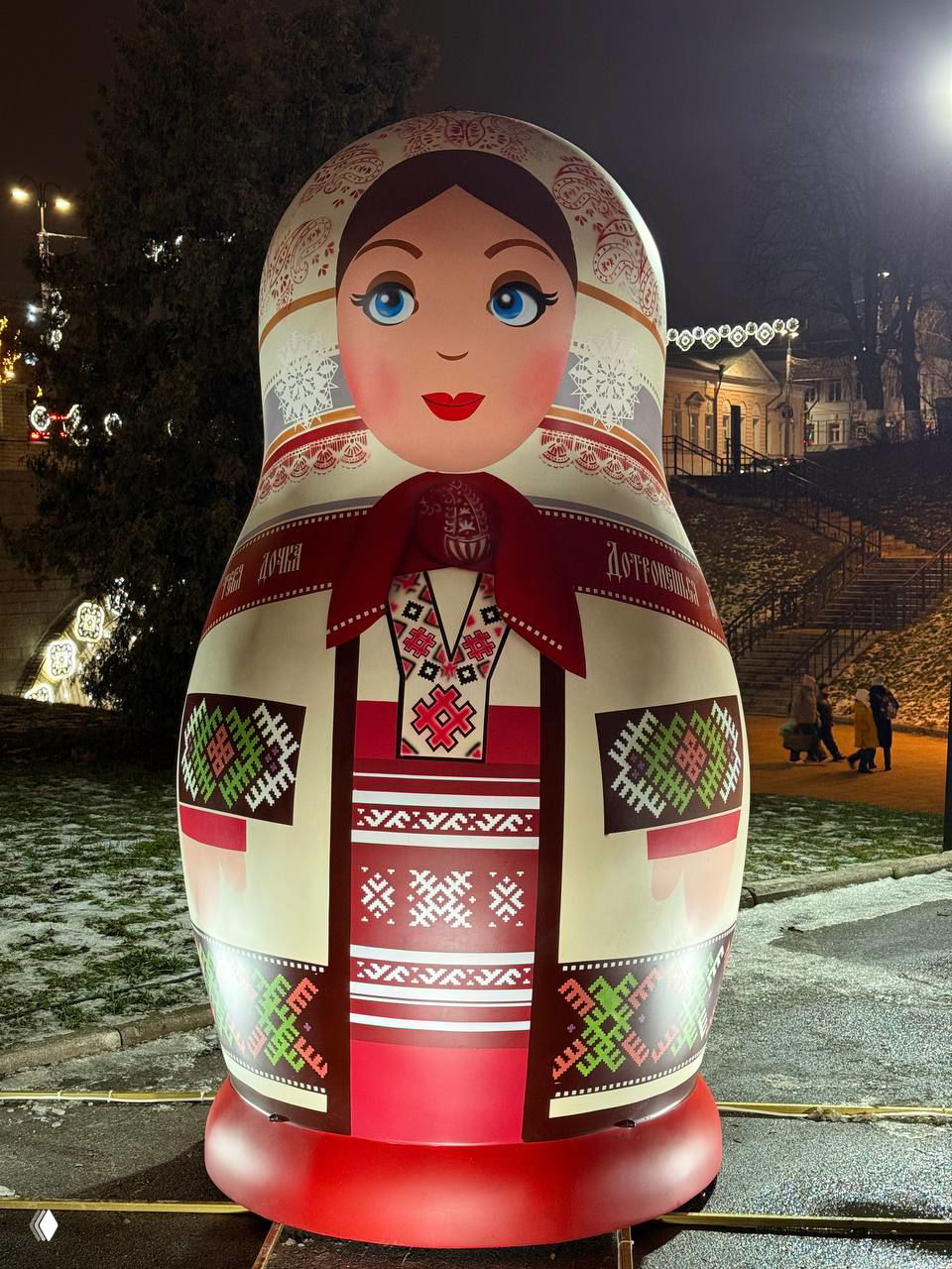 Матрешка в светло-кремовом и красном народном костюме на ночной прогулочной зоне Лыбедского бульвара, рядом видна архитектура города.
