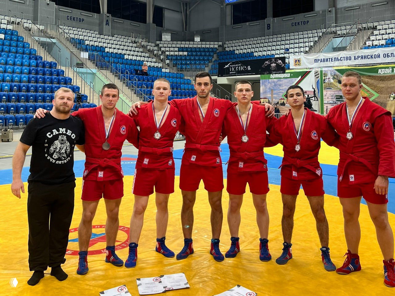 Командное фото рязанских самбистов в красных кимоно с медалями на воротнике на ковре в спортзале Брянска после турнира.