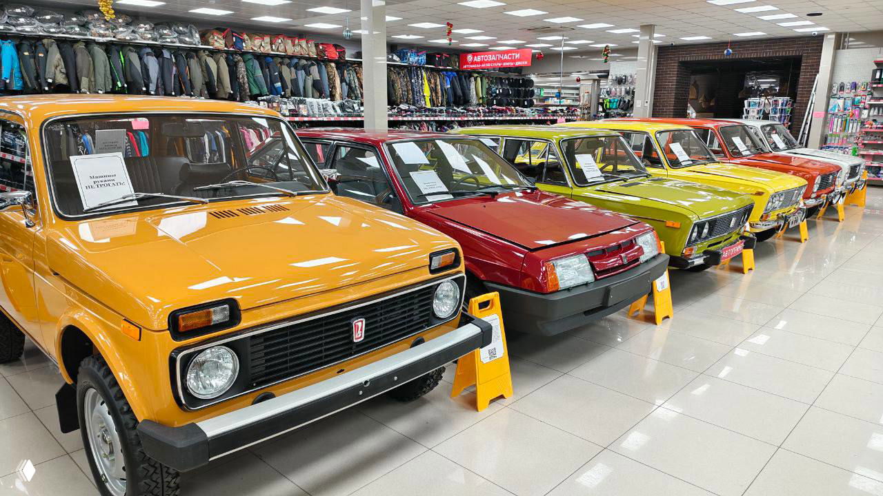 Ряд советских внедорожников и легковых машин, включая жёлтую Ниву и красные ВАЗы в экспозиции, выставочный зал Горроща, посетители и указатели за машинами, Рязань, ул. Островского.