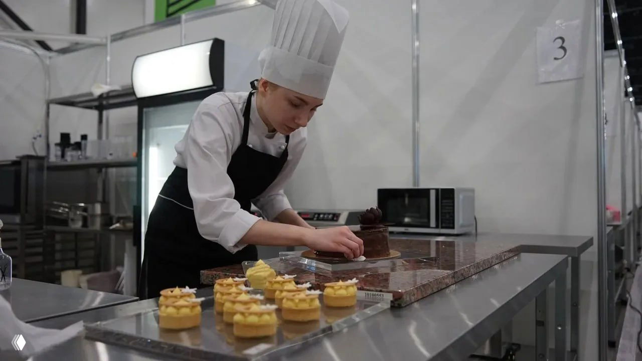 Студентка в поварском колпаке украшает десерты на рабочем столе в зоне «Кондитерское дело» на чемпионате «Профессионалы»