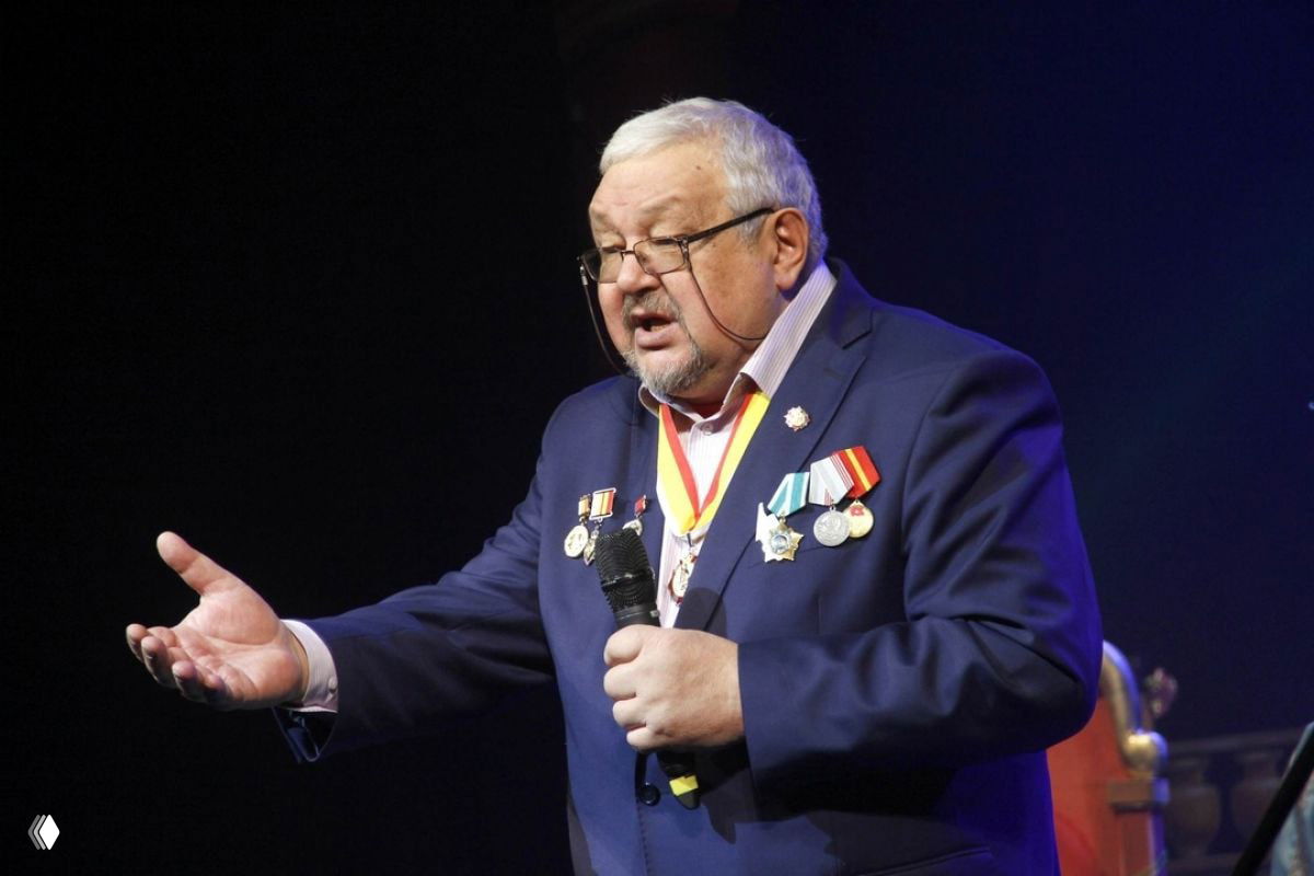 Валерий Шадский на сцене в синем костюме с медалями и орденами, держит микрофон во время выступления, средний план