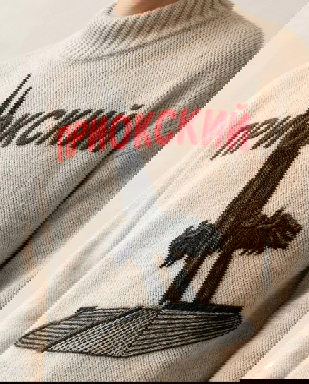 Тёплый светлый свитер с надписью «ПРИОКСКИЙ» и вышитым памятником со взлетающими вертикальными элементами на постаменте, крупный план работы дизайнера.