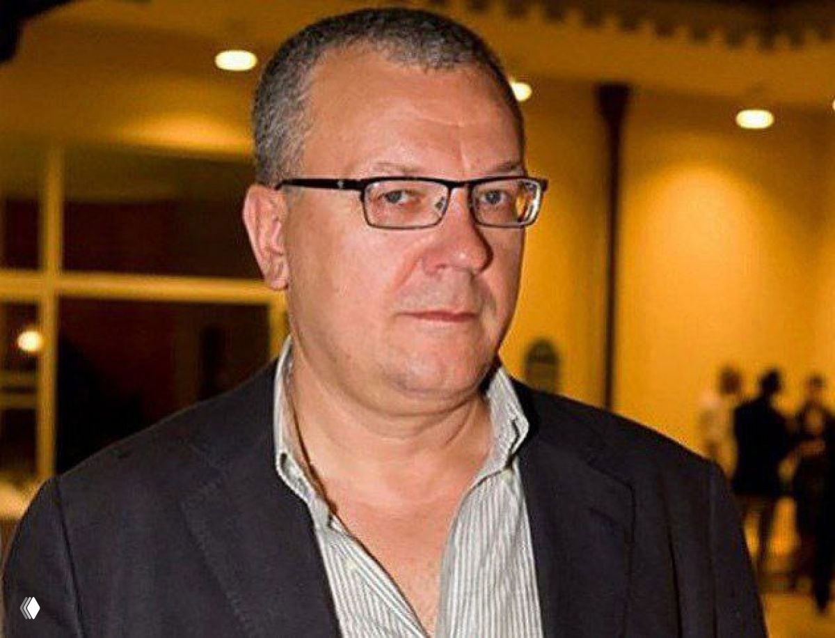 Портрет мужчины средних лет в очках и пиджаке на фоне внутреннего пространства с тёплым освещением; деловой портрет.