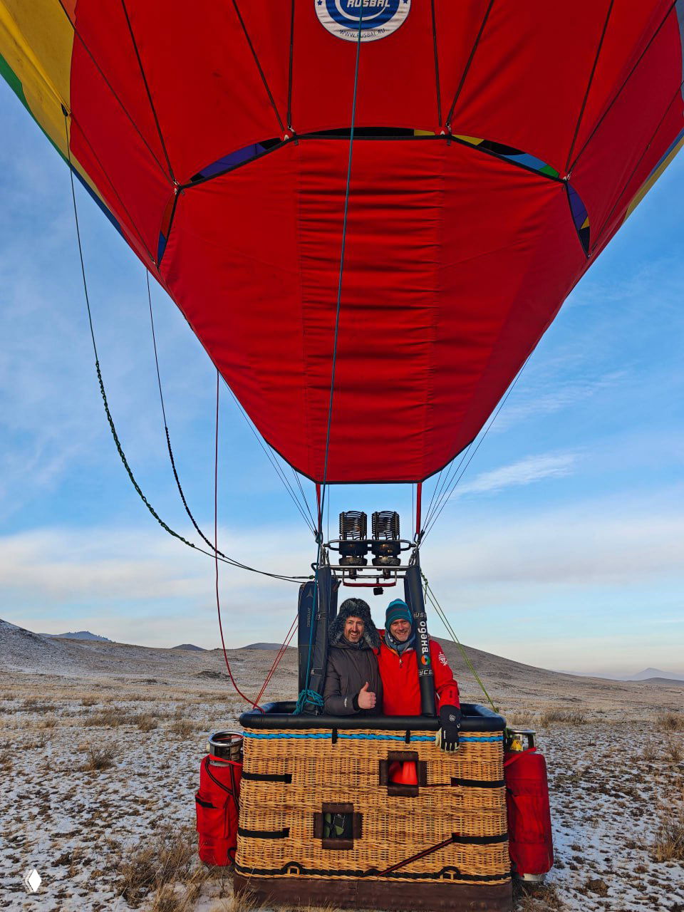 Пилоты в плетёной корзине красного аэростата, вид на купол сверху; снежная равнина и бескрайняя сибирская тайга вокруг.