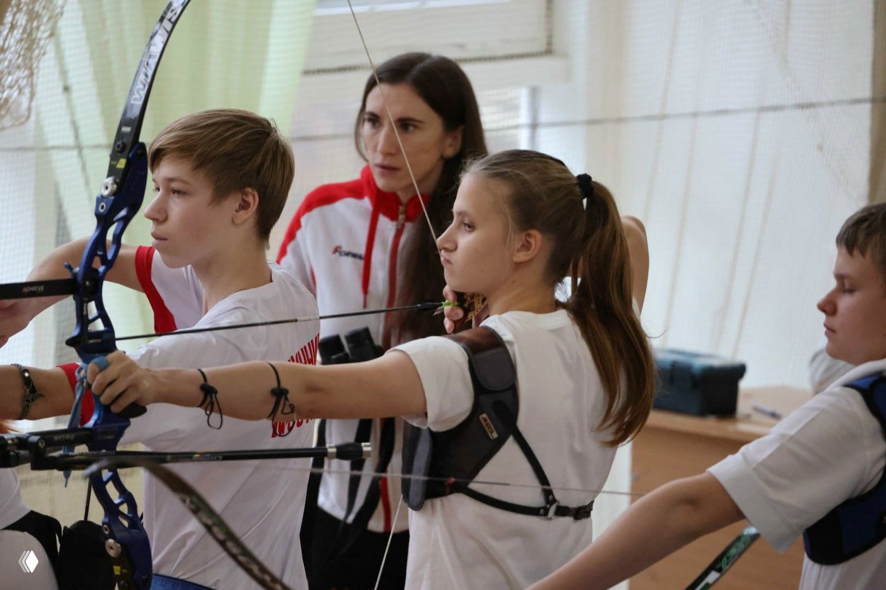 Юная лучница при выстреле из классического лука в спортивном комплексе «Буревестник», рядом тренер и другие участники соревнований.
