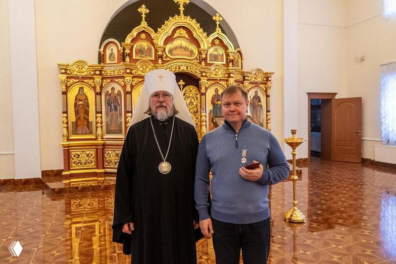 Митрополит Марк позирует вместе с депутатом Олегом Владимировичем Шишовым, держит медаль II степени на фоне декоративного иконостаса в храмовом зале.