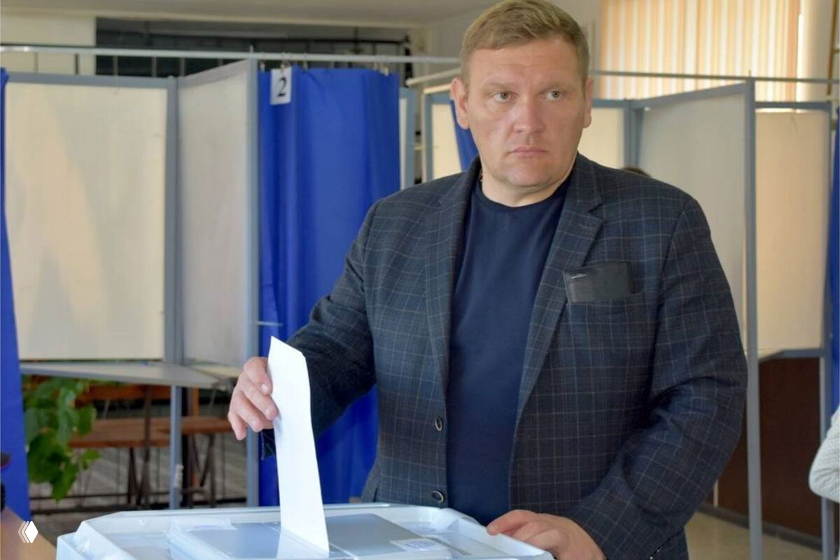 Мужчина в клетчатом пиджаке опускает бумажный бюллетень в прозрачную урну для голосования на участке с кабинками и ширмами.