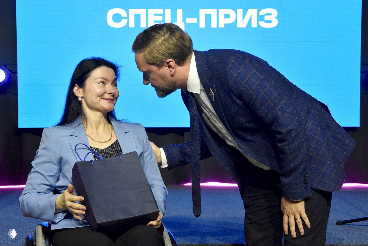 Женщина в инвалидной коляске принимает подарок на сцене церемонии награждения; рядом мужчина вручает пакет, на фоне экран с надписью «СПЕЦ-ПРИЗ».