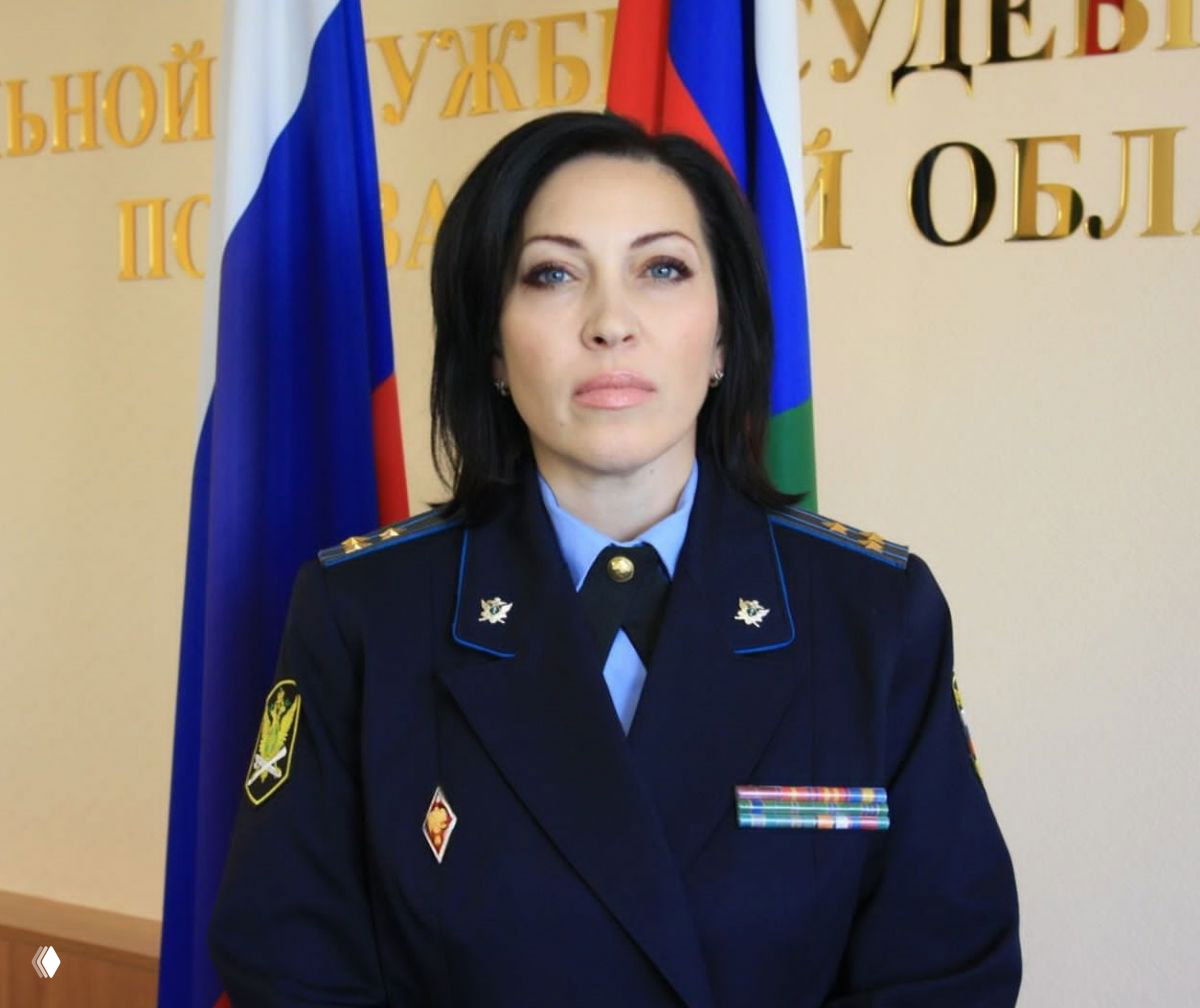 Портрет женщины в форменной одежде судебного пристава на фоне флагов и стенда Управления Федеральной службы судебных приставов по региону.