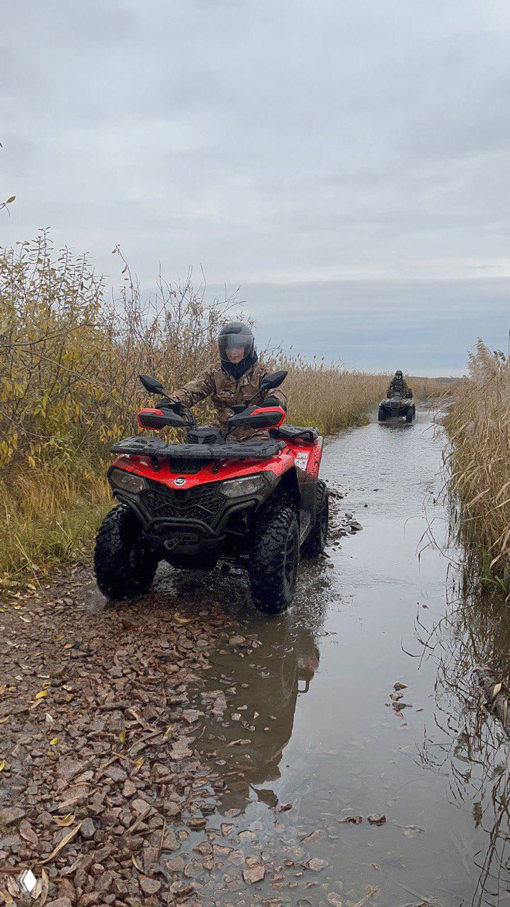Полноприводный квадроцикл на лесной тропе и лужах, водитель в шлеме и экипировке проезжает через болотистый участок маршрута проката в Челябинске; пейзаж осенний.