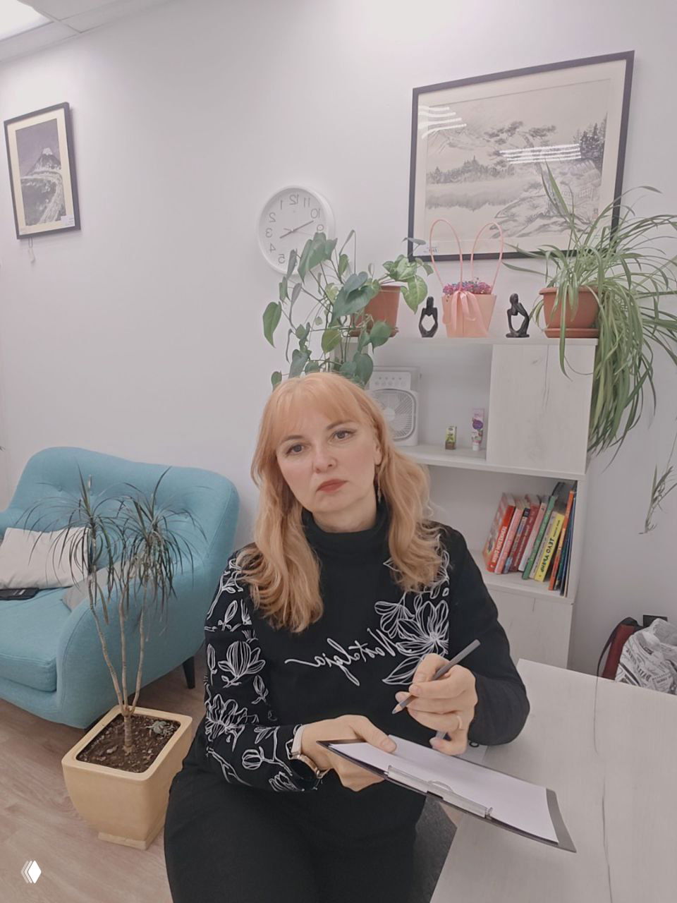 Женщина-психолог сидит в кабинете с блокнотом и ручкой, вокруг — комнатные растения, кресло и книжная полка на заднем плане.