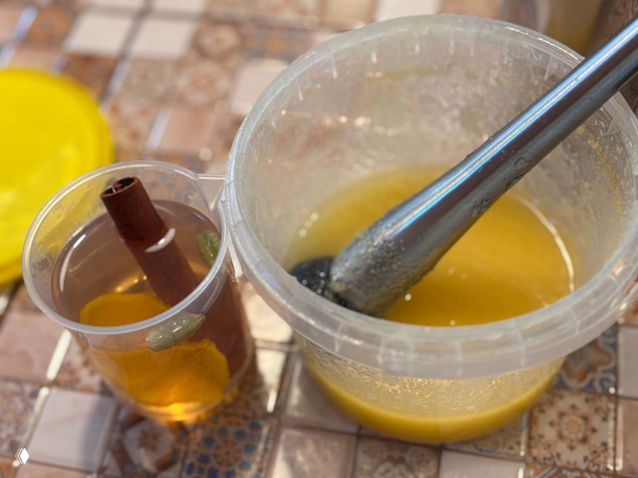 Пластиковая ёмкость с медовой массой и погружным блендером, рядом стакан с настоем пряностей: палочка корицы, цедра лимона и кардамон.