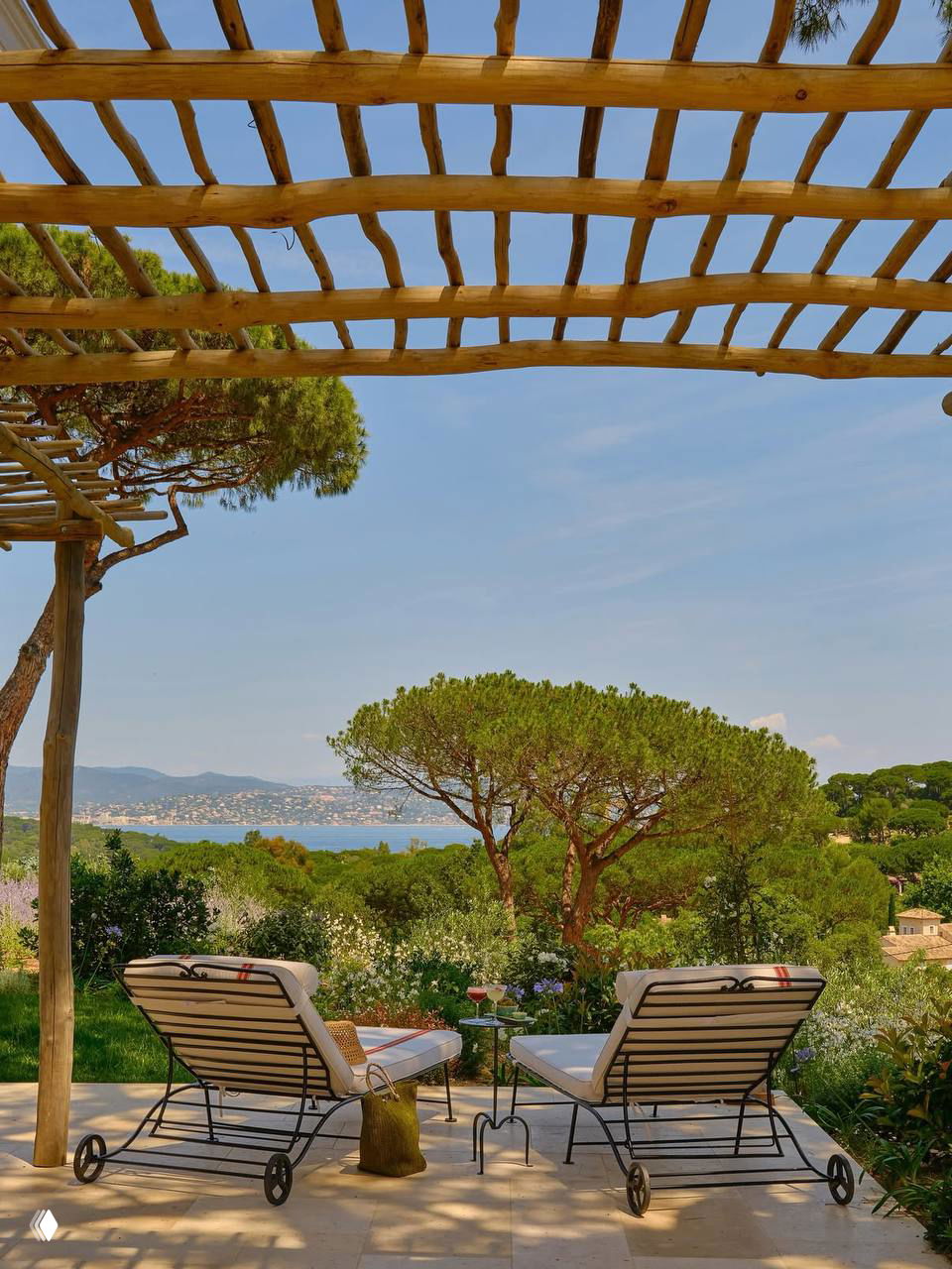 Зона отдыха с шезлонгами под перголой и видом на залив: шезлонги, столик, зелень и спокойное море на горизонте, типичный курортный пейзаж.
