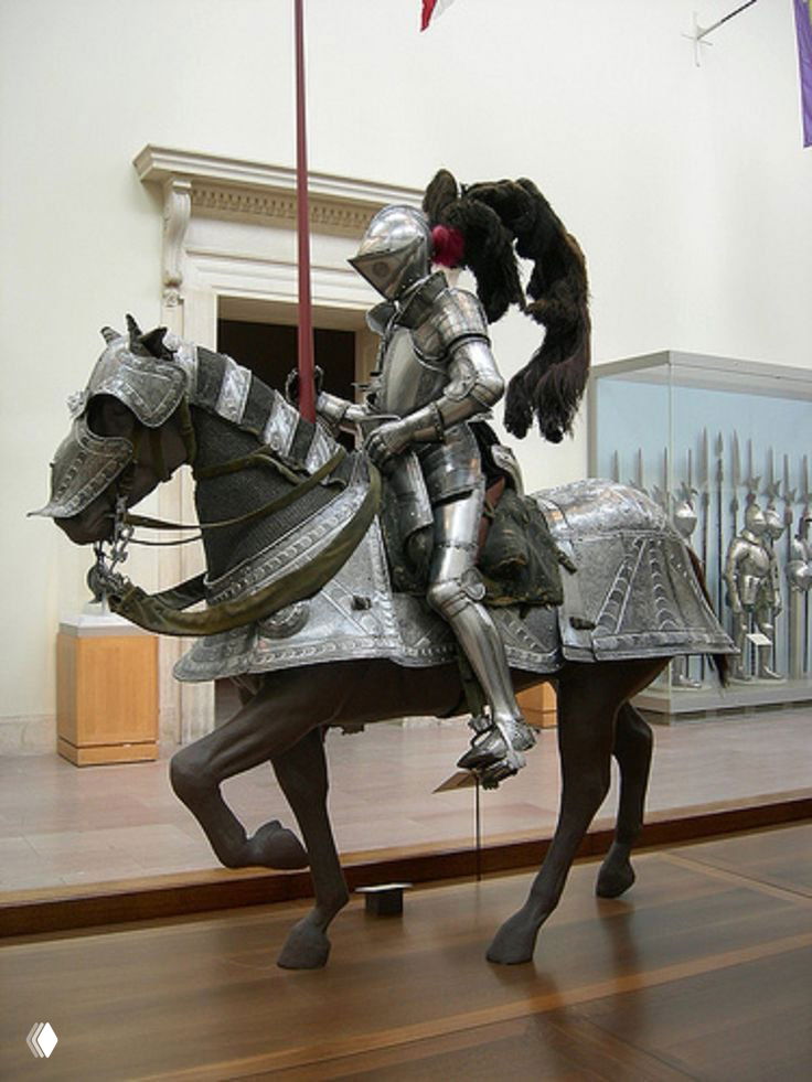Манекен всадника в полном стальном рыцарском доспехе вместе с бронированным конём в музейной витрине, вид в профиль.