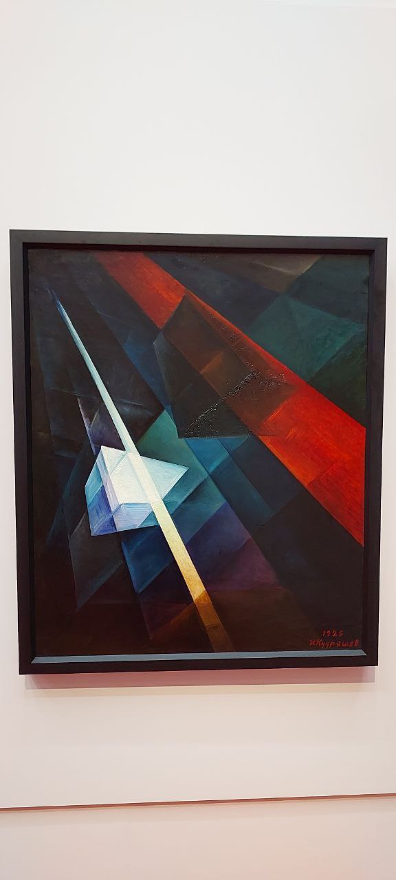 Фотография абстрактной геометрической картины с насыщенными красно-синими и тёмно-зелёными плоскостями, висящей в музейном зале.