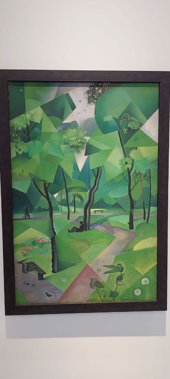 Фото полотна с модернистским зелёным пейзажем, собранным из геометрических форм и плоскостей, выставленного на белой стене галереи.