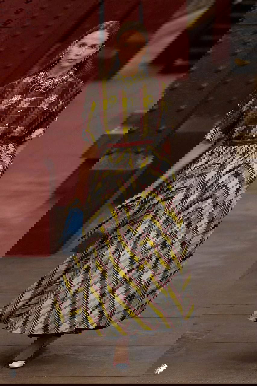 Модель в клетчатой юбке и рубашке идёт по платформе метро Chanel; мощные колонны и скамьи создают сценографию показа Métiers d’art, Маттье Блэзи.