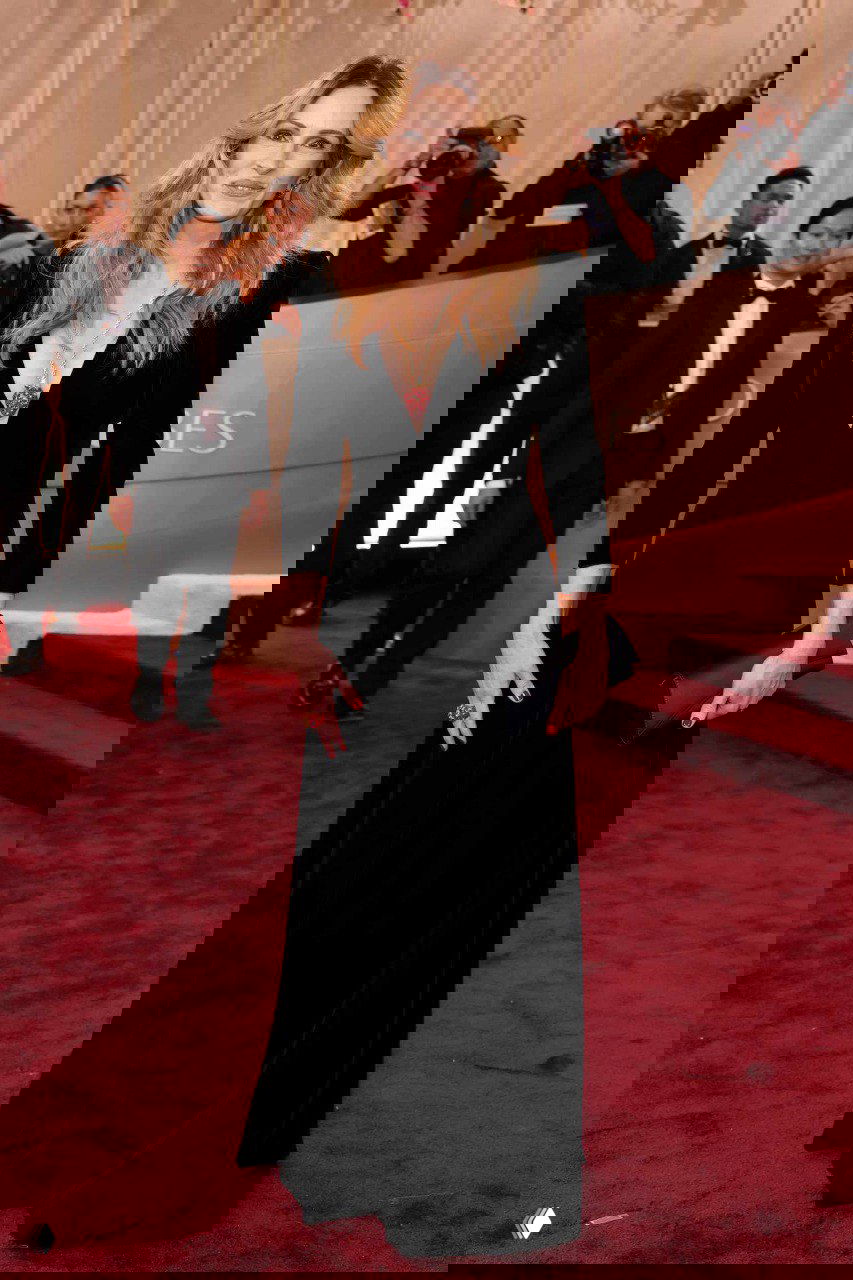 Женщина на красной дорожке в элегантном чёрном платье с кулоном в форме клубники; позади фотографы и гости церемонии.