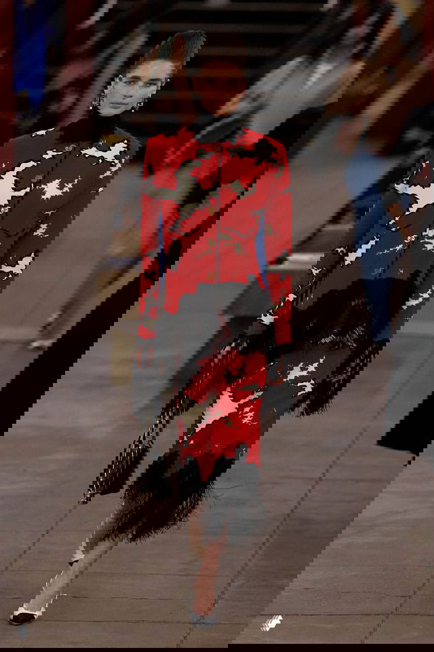 Модель в красном комплекте с бахромой идёт по подиуму-станции метро; детали и обувь с контрастным носком подчёркивают идеи коллекции Métiers d’art.