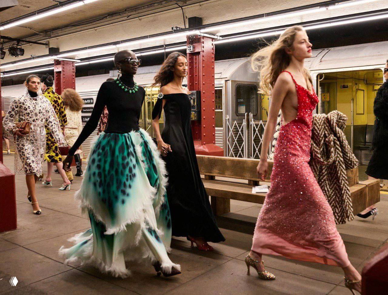 Группа моделей в вечерних платьях и объемных юбках проходит по платформе, создавая контраст между высокой модой и повседневной городской обстановкой метро.