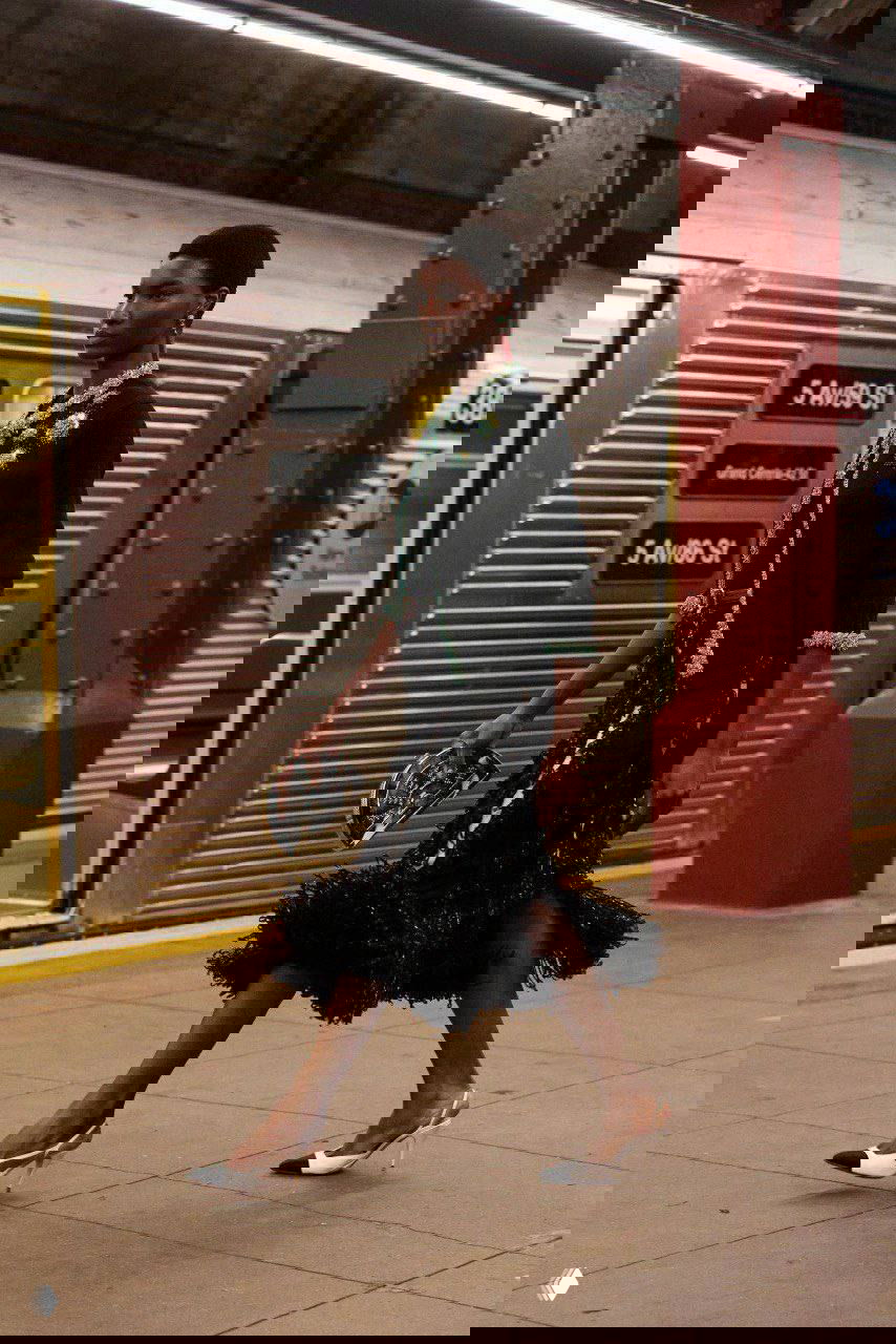 Темнокожая модель в черном костюме с текстурной юбкой и украшениями идет по платформе рядом с металлическим вагоном метро во время шоу Chanel.