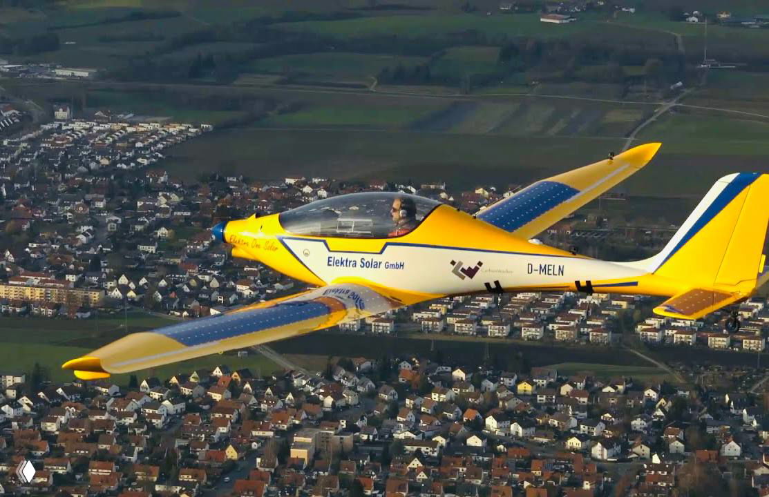 Жёлтый одноместный самолёт Elektra Solar в полёте над поселением, виден пилот и солнечные панели на крыльях, демонстрация электрической авиации.