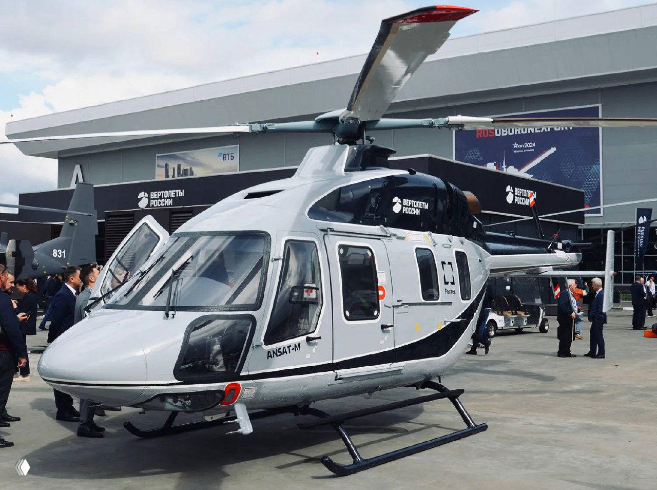 Вертолёт Ансат‑М на стоянке авиасалона с логотипами «Вертолёты России» и Ростеха, люди возле экспозиции и демонстрационные стенды.