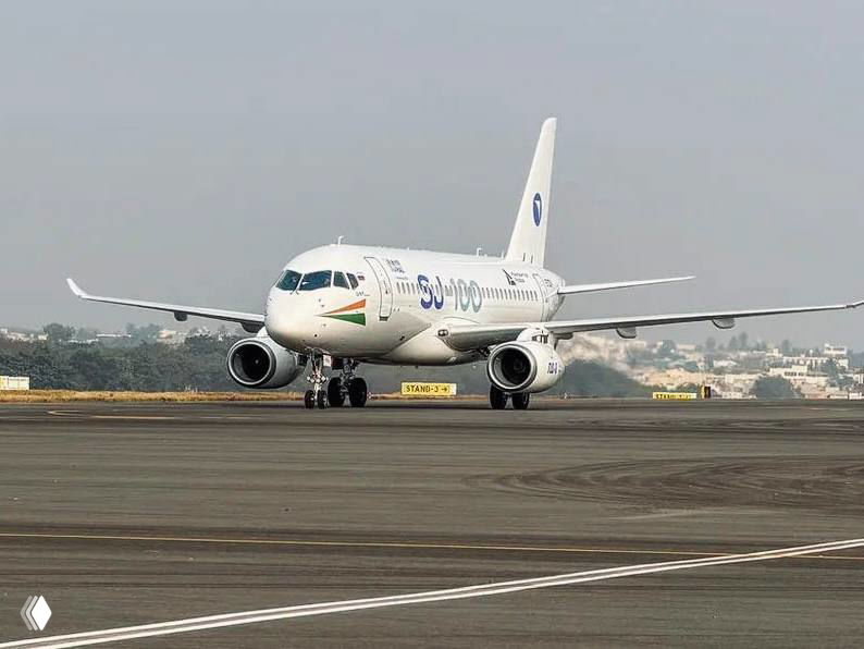 Superjet (SJ) на рулёжной дорожке в Хайдарабаде: вид спереди белого лайнера с ливреей и логотипом, самолёт на фоне аэродрома во время выставки.