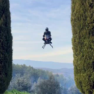 Фото: пилот на Volonaut Airbike зависает между высокими деревьями на фоне холмов, аппаратура и посадочные опоры чётко видны.