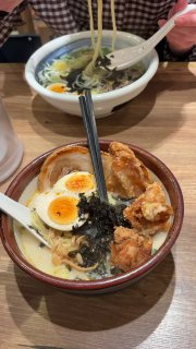 Близкий план миски рамена с бульоном, яйцом и кусочками жареного мяса: человек поднимает лапшу палочками, видны гарниры и текстура бульона.