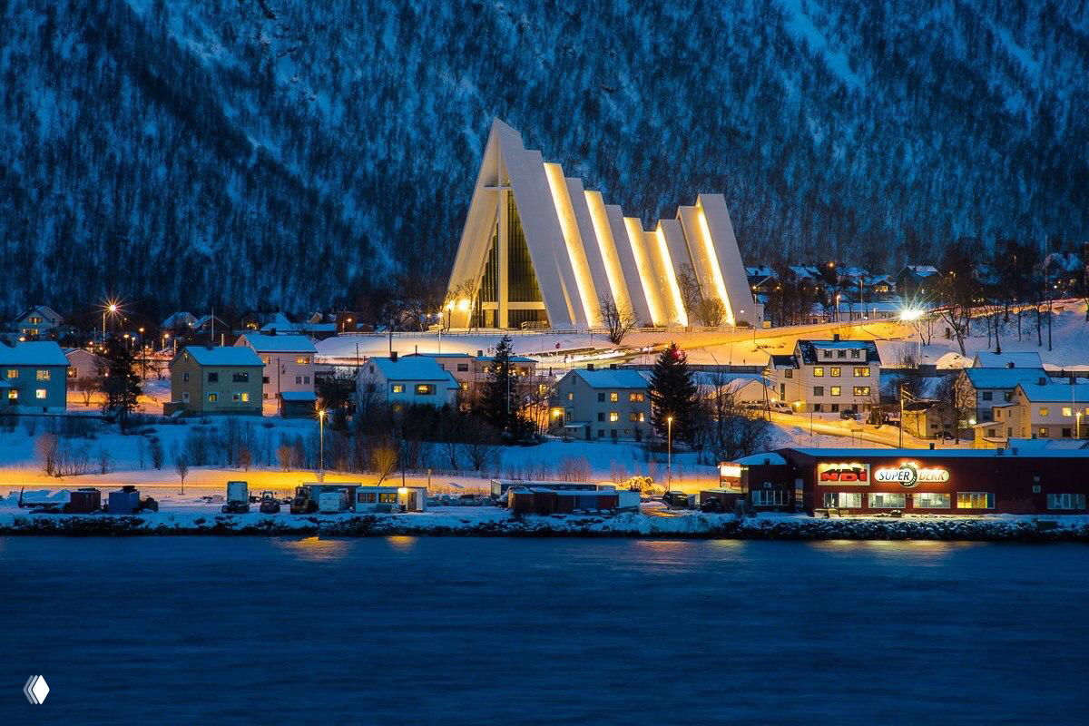 Ночная фотография прибрежного города с освещённым современным зданием (возможно собор) у воды, снежные склоны на заднем плане.