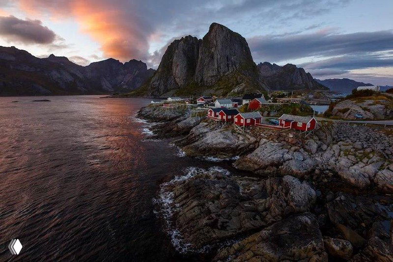 Норвежский прибрежный рыбацкий поселок с красными домиками на скалистом берегу и высокими горами — спокойный закатный пейзаж фьорда.