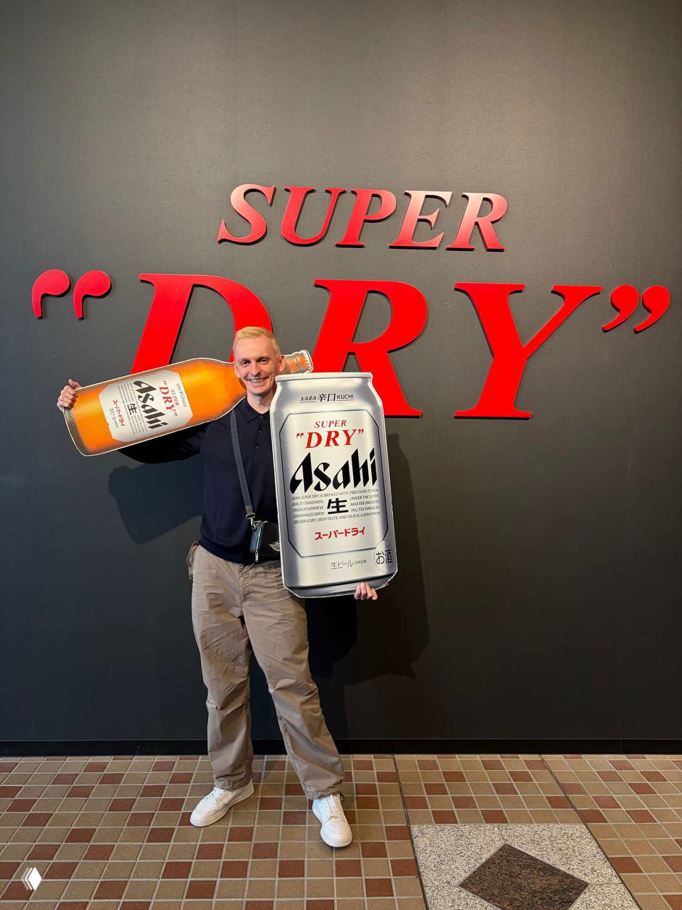 Посетитель фотографируется у бренд-стены Super "DRY", держа увеличенные реквизиты — банку и бутылку Asahi, фотозона для гостей.