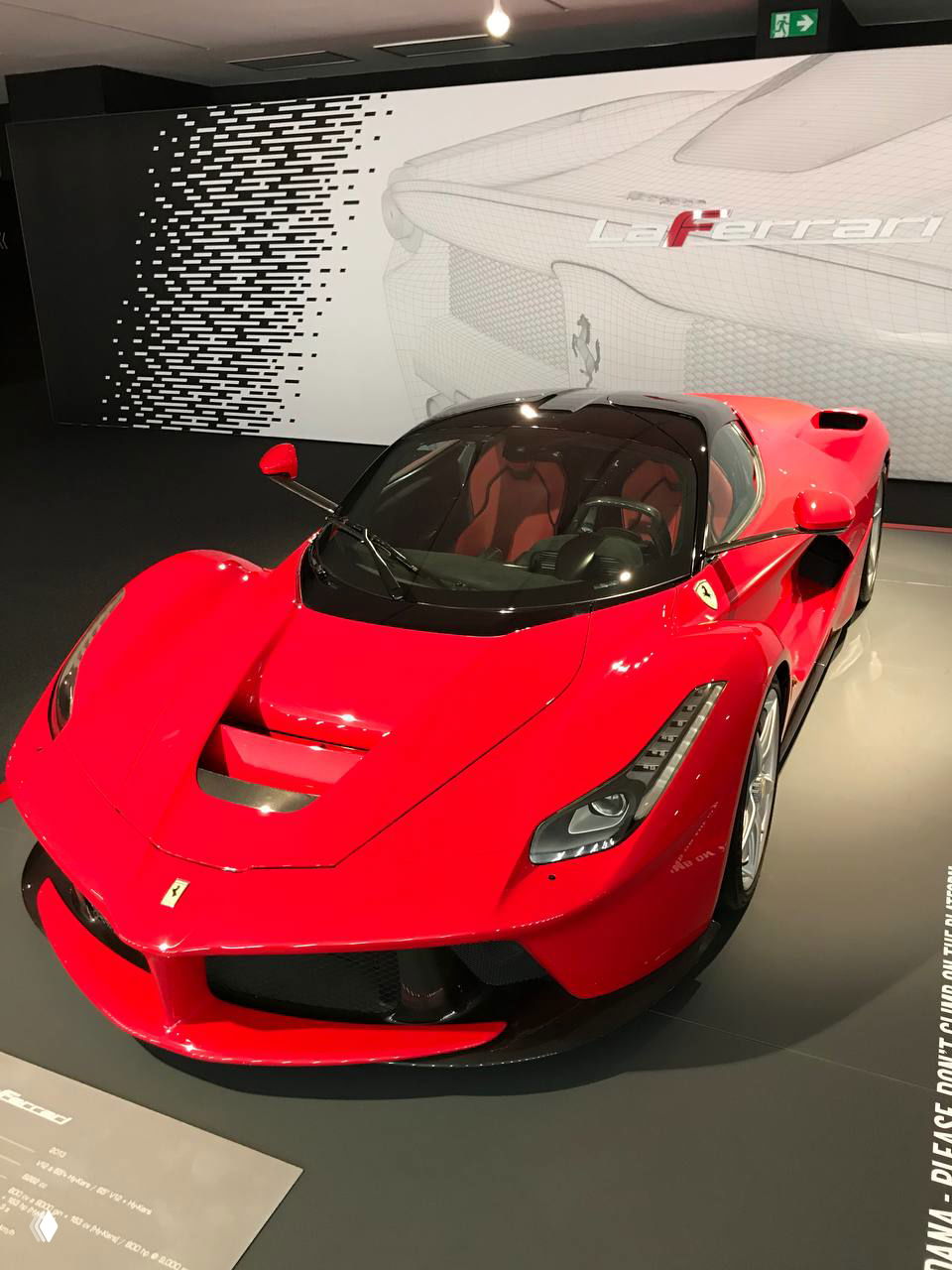 Ярко-красная Ferrari на демонстрационном подиуме в тёмном зале с оформлением стен и логотипом марки позади автомобиля.